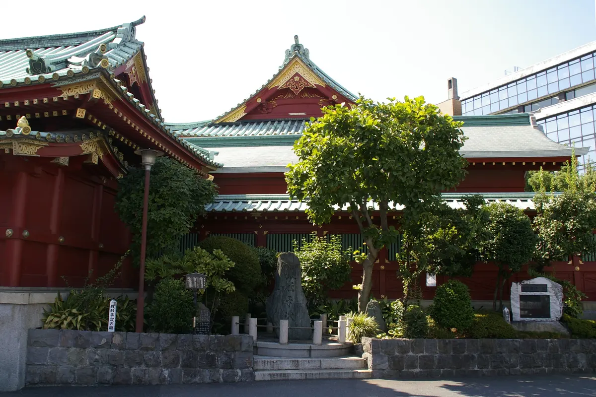 Kanda Myojin'de ağaçların arkasında yeşil çatılı vermilyon bir tapınak binası ve alçak bir duvarın dibinde taş bir anıt